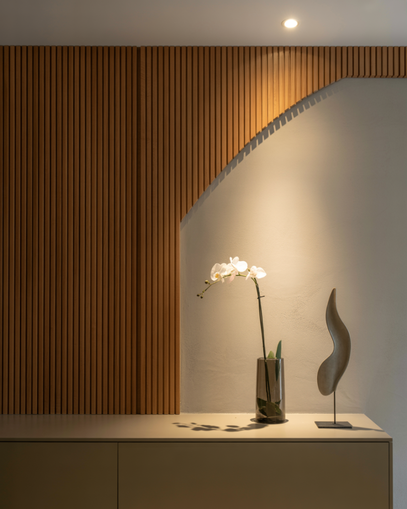 Minimalist entryway with modern slatted wood wall paneling and a curved cutout detail, softly lit by a recessed ceiling light. A sleek console below holds a single white orchid in a glass vase and an abstract metal sculpture. Elegant and serene decor with warm architectural accents