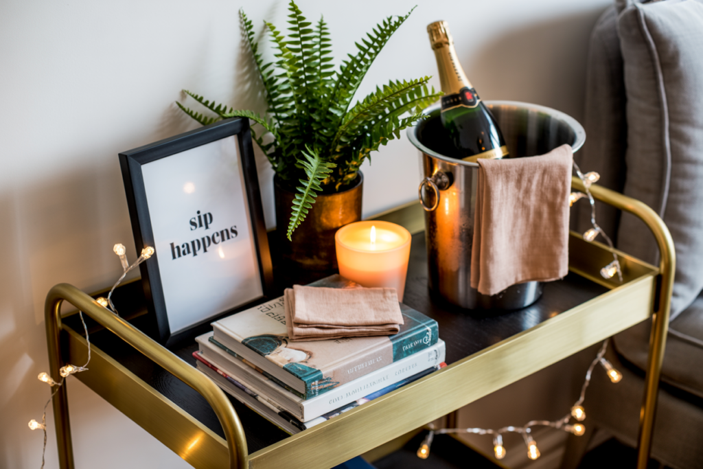 Cozy and playful bar cart styling with a framed quote that reads 'sip happens,' a potted fern, champagne bottle in a silver ice bucket, glowing candle, neutral cloth napkins, stacked books, and string lights draped around a gold and black bar cart. Set beside a gray armchair for a warm, inviting vibe.