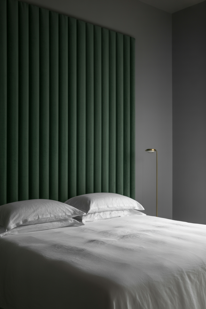 Minimalist bedroom with a dramatic floor-to-ceiling emerald green vertical velvet headboard, crisp white bedding with two stacked pillows, and a sleek brass floor lamp against a matte gray wall.
