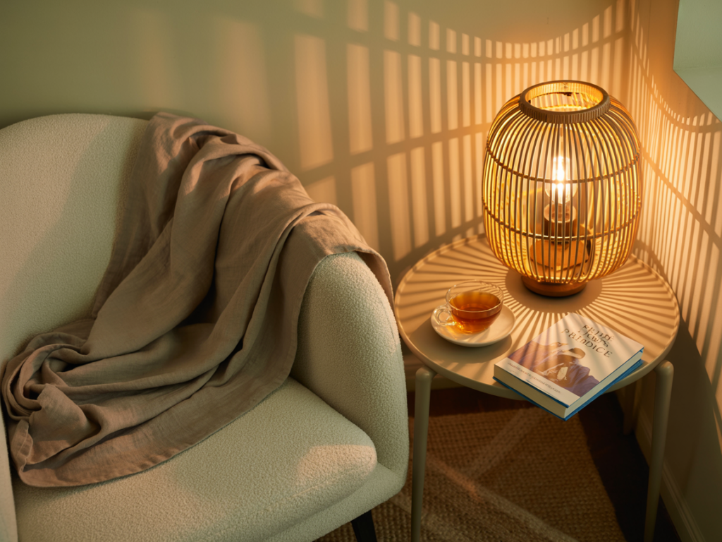 A cozy reading corner lit by a glowing rattan lamp casting patterned shadows on the wall. A boucle armchair with a soft taupe throw sits beside a small round side table. On the table are a glass cup of tea and a copy of Pride and Prejudice. A jute rug lies underneath, completing the warm, inviting, and serene atmosphere.