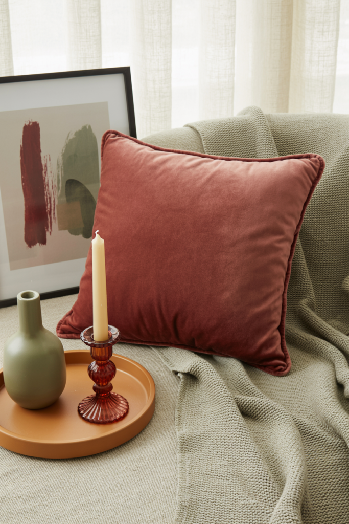 "Cozy fall decor vignette with cranberry velvet pillow, sage green knit throw, caramel tray with green vase and red glass candleholder, styled by a window with soft neutral curtains and abstract wall art